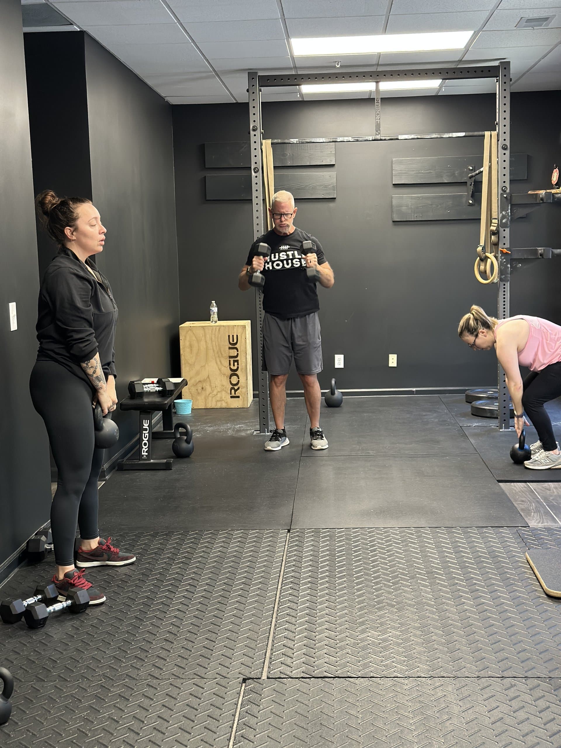 Members working with kettlebells and dumbbells beside the squat rack at Hustle House Health & Wellness in Woodstock, GA