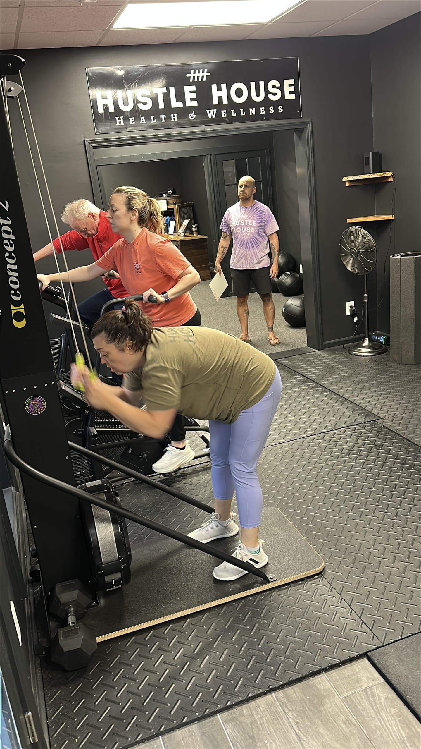 Members on the rowing machines under the Hustle House Health & Wellness sign in Woodstock, GA
